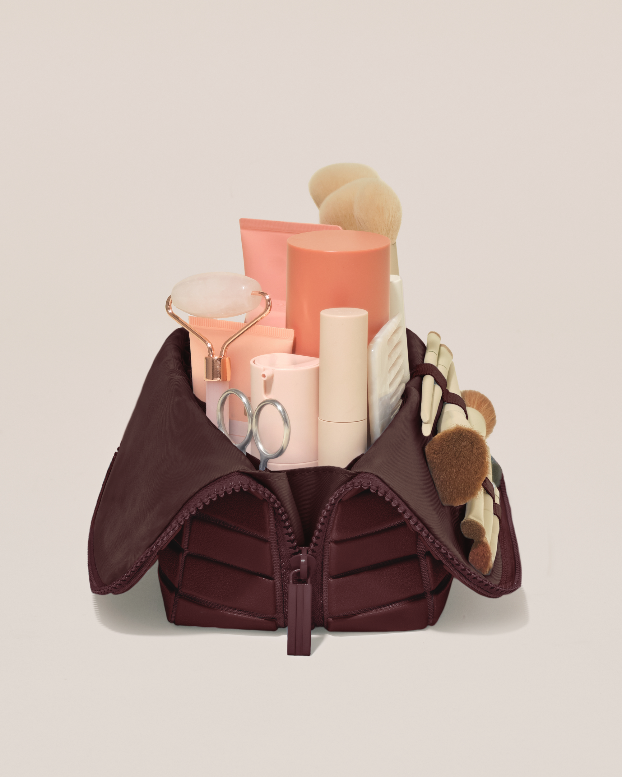Side profile of the Medium Cassis (maroon) Traveller makeup bag filled with beauty essentials standing upright, showing flexible compartment design