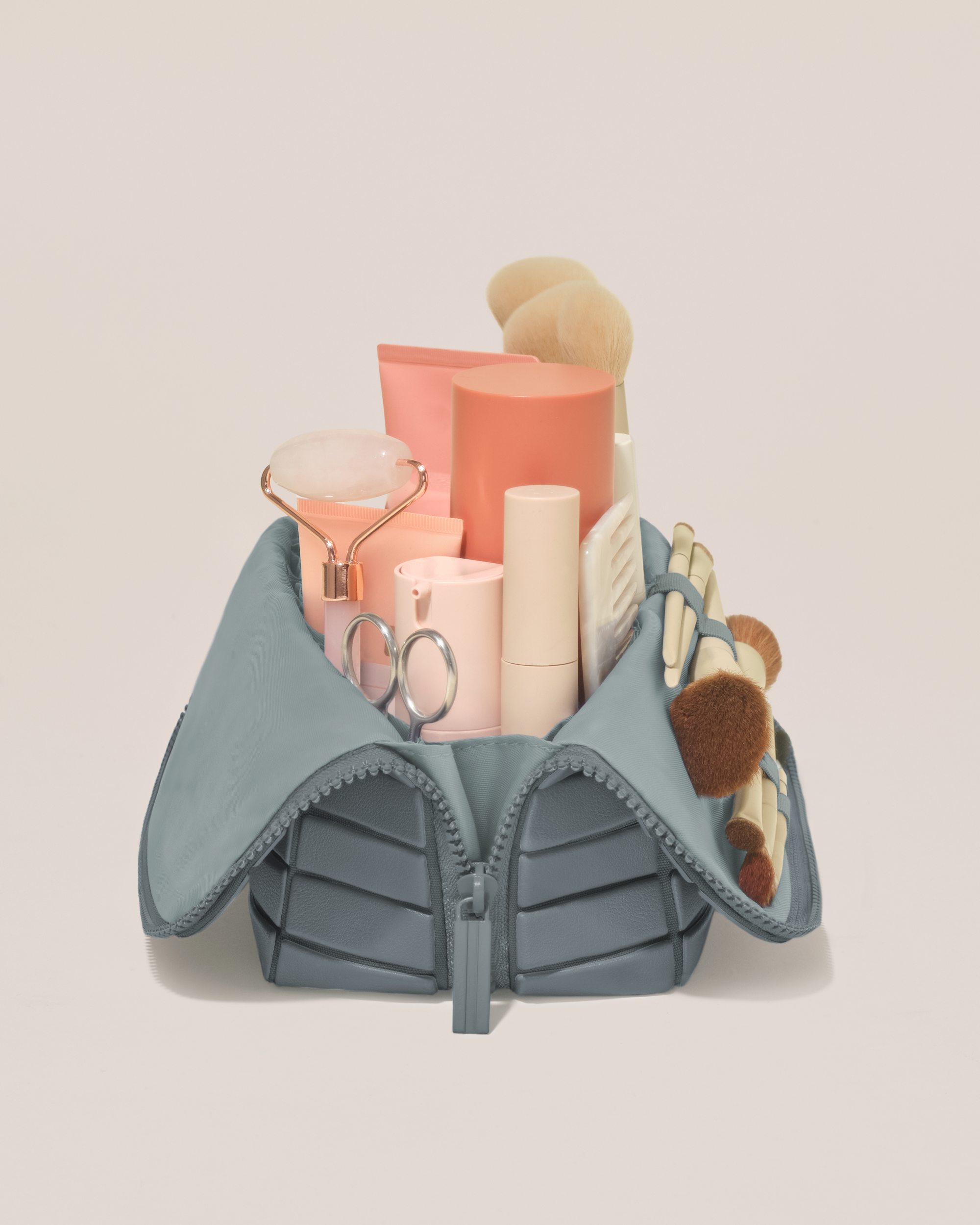 Side profile of the Medium Slate (blue) Traveller makeup bag filled with beauty essentials standing upright, showing flexible compartment design